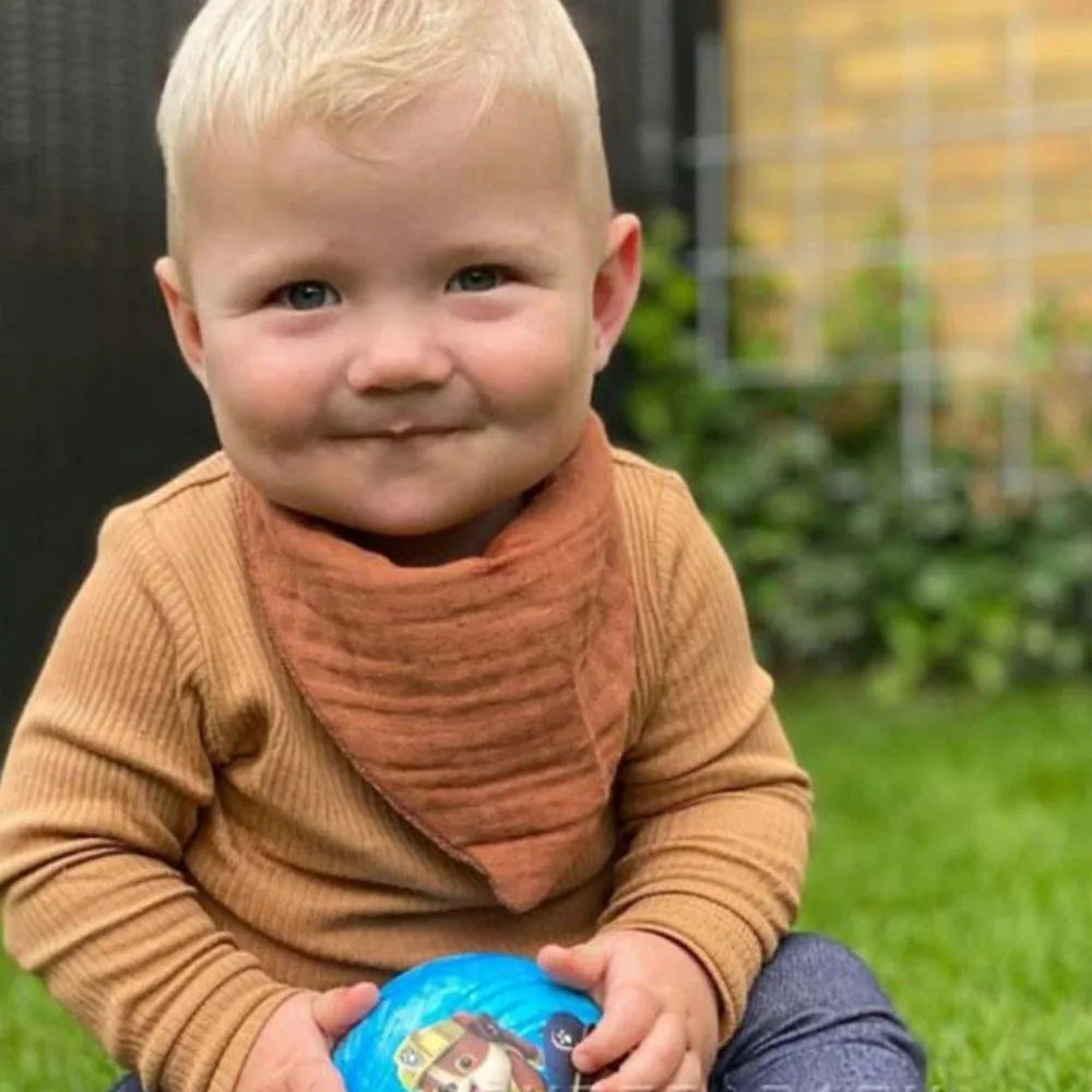Un tout-petit joyeux avec un sourire éclatant, portant des accessoires douillets BABY PREMA, dont un pull marron et une écharpe, assis sur l'herbe et tenant de manière ludique un ballon coloré.