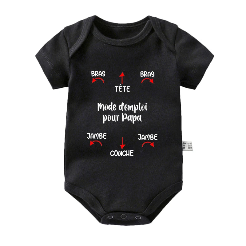 Une combinaison bébé noire avec texte blanc et rouge en français, parfaite pour les journées chaudes. Le texte comprend des flèches pointant vers différentes parties de la barboteuse en coton avec des étiquettes : « Bras » (Bras), « Tête » (Tête), « Jambe » (Jambe) et « Couche » (Couche). Le texte central indique "Mode d'emploi pour Papa". Il s'agit du Body Coton Imprimé de BABY-PREMA.