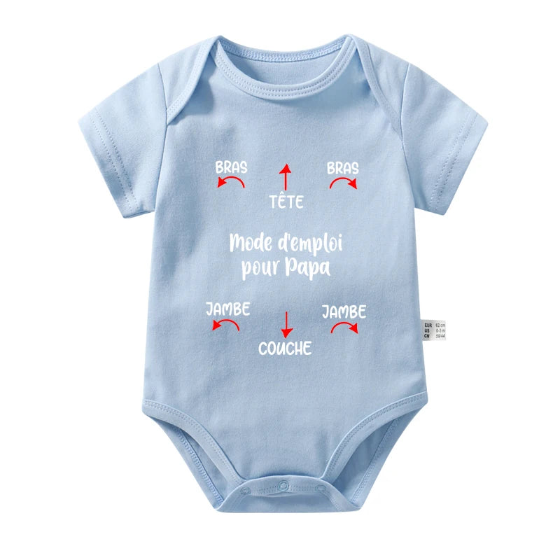 Un Body Coton Imprimé bleu clair de BABY-PREMA est présenté avec des instructions humoristiques en français pour habiller le bébé. Les flèches pointent vers chaque manche intitulée « Bras », le décolleté intitulé « Tête », le bas intitulé « Jambe » et la zone de la couche intitulée « Couche ». Le texte dit « Mode d'emploi pour Papa ». Parfait pour les journées chaudes.