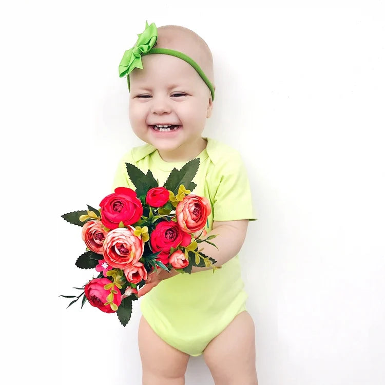 Un bébé dans un Lot de 7 Bodies Bébé Imprimés Unis jaune de BABY-PREMA avec un bandeau vert tient un bouquet de fleurs rouges et roses, souriant brillamment sur un fond blanc.