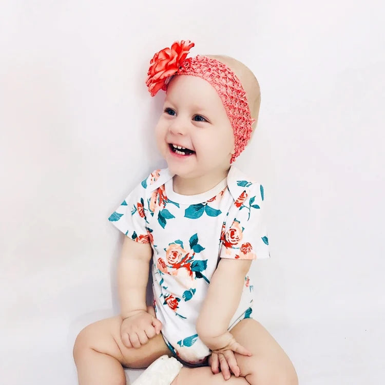 Un bébé souriant portant un Lot de 7 Bodies Bébé Imprimés Unis de BABY-PREMA fleuri et un bandeau rose avec une fleur est posé sur un fond uni.