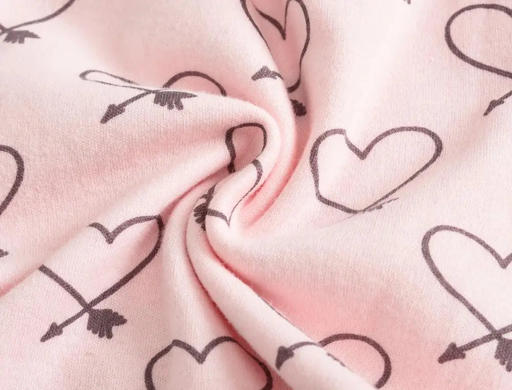 Un gros plan d'un tissu en coton rose doux avec un motif de cœur et de flèches noirs, parfait pour le Lot de 7 Bodies Bébé Imprimés Unis de BABY-PREMA. La matière est légèrement froissée au centre.