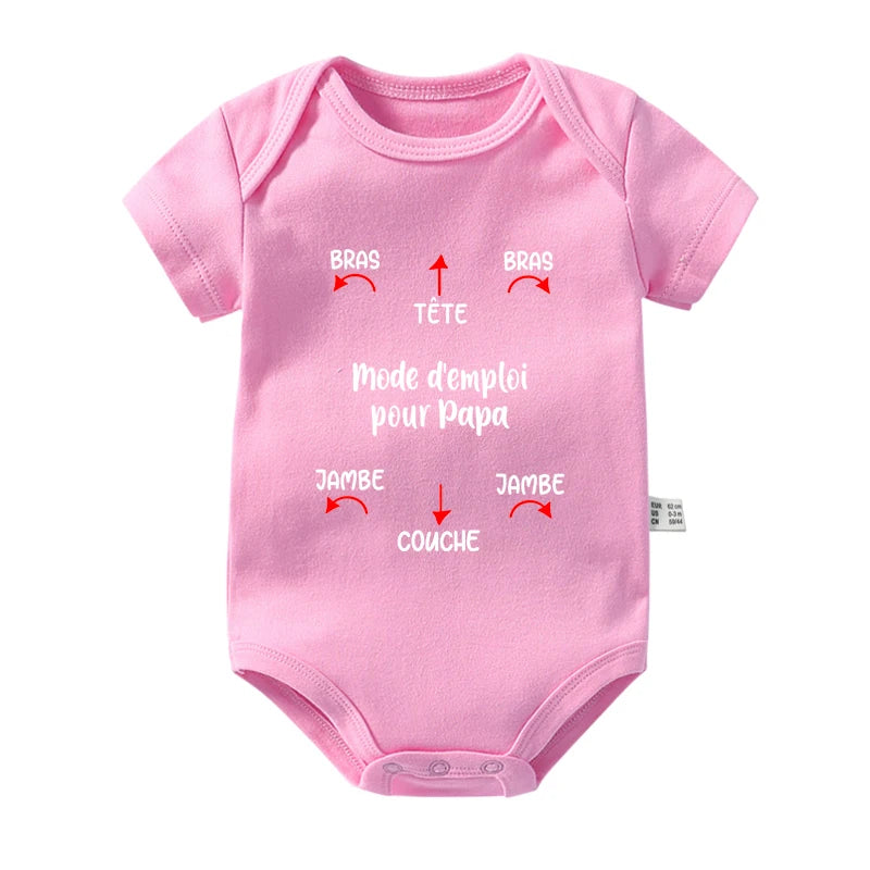 Une combinaison bébé en coton rose, parfaite pour les journées chaudes, comporte des instructions en français indiquant « Mode d'emploi pour Papa ». Les flèches pointent vers différentes zones intitulées « Bras » (bras), « Jambe » (jambe), « Tête » (tête) et « Couche » (couche). Ce charmant Body BABY-PREMA en Coton Imprimé possède des manches courtes et des fermetures à pression dans le bas.