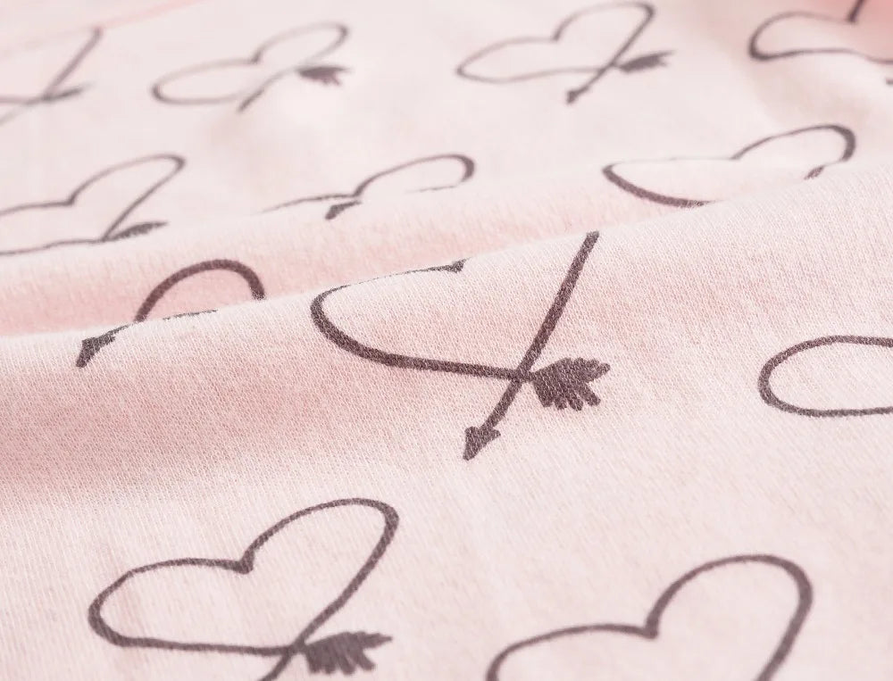 Un gros plan d'un tissu en coton rose avec un motif imprimé de cœurs et de flèches noirs, couramment utilisé pour le Lot de 7 Bodies Bébé Imprimés Unis de BABY-PREMA, avec des boutons à pression pratiques.
