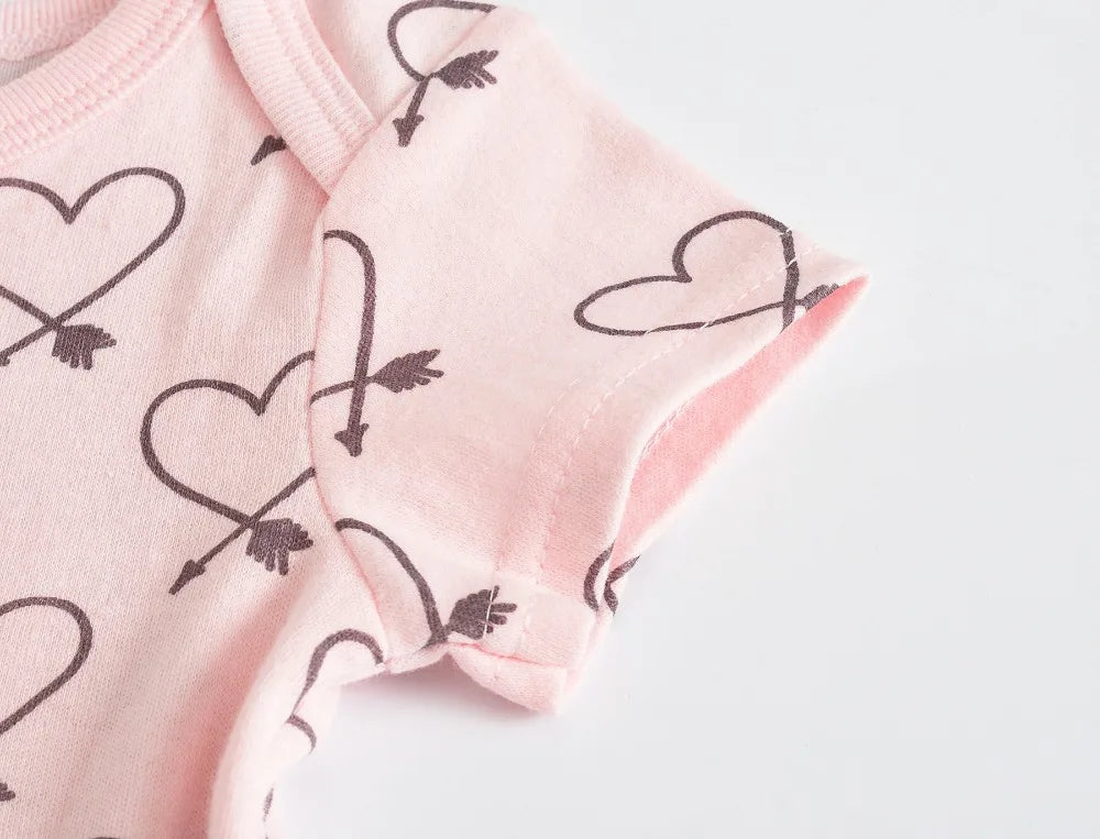 Gros plan d'une combinaison rose pour bébé en coton doux avec des motifs de cœurs et de flèches noirs, avec des boutons pratiques à pression sur un fond blanc. Produit : Lot de 7 Bodies Bébé Imprimés Unis par BABY-PREMA.
