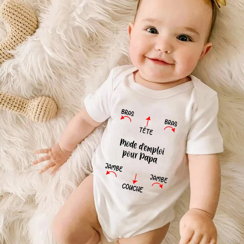 Un bébé portant un Body Coton Imprimé blanc de BABY-PREMA avec du texte français et des flèches en noir et rouge imprimés dessus. Le texte indique « Mode d'emploi pour papa » et les flèches pointent vers différentes parties du corps (tête, bras, jambes, couche) étiquetées en français. Le bébé porte un bandeau à nœud et est allongé sur une couverture douce et de couleur claire.