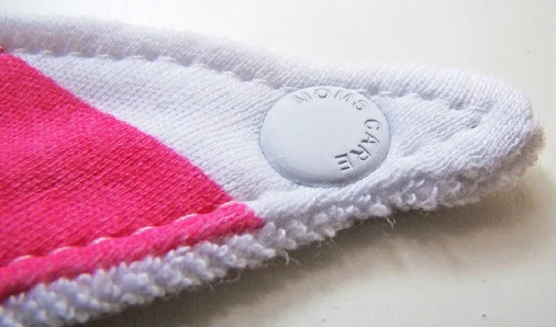 Gros plan sur un article en tissu blanc et rose avec un bouton pression rond blanc. Les points de suture sont visibles et le bouton-pression est gravé des mots « MOM’S CARE ». Le Lot de 3 pièces Bavoirs Bébé de BABY-PREMA offre une texture douce semblable à une serviette sur un bord.