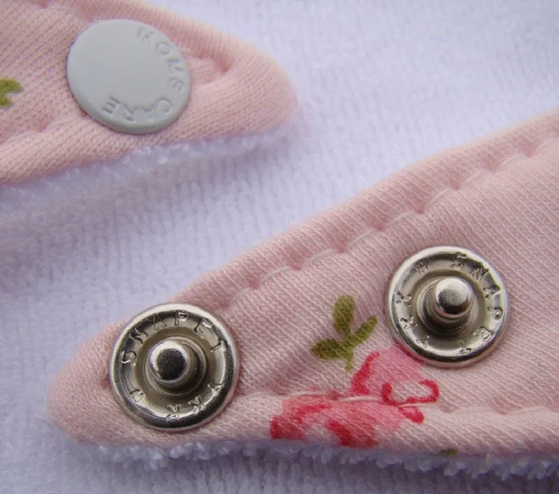 Gros plan d'un tissu rose avec des motifs floraux et des boutons-pression en métal attachés à un vêtement blanc. Les boutons ronds portent un texte gravé et sont solidement fixés, affichant les textes « SNAP » et « CARE ». Fabriqué à partir de coton de haute qualité, le tissu présente un point zigzag le long du bord. Produit phare : Lot de 3 pièces Bavoirs Bébé de BABY-PREMA.
