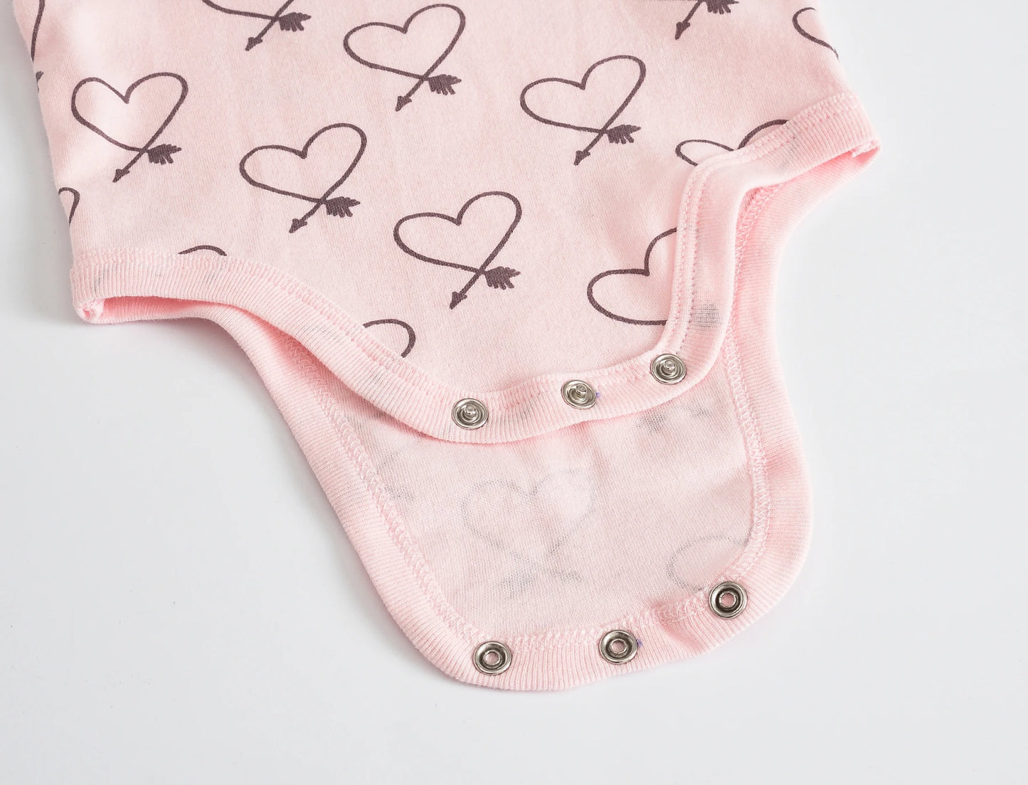 Gros plan d'une combinaison rose en coton doux présentant un motif cœur et flèche, avec des fermetures à pression en bas pour une fermeture facile. Ce pratique Lot de 7 Bodies Bébé Imprimés Unis de BABY-PREMA est parfait à porter au quotidien.