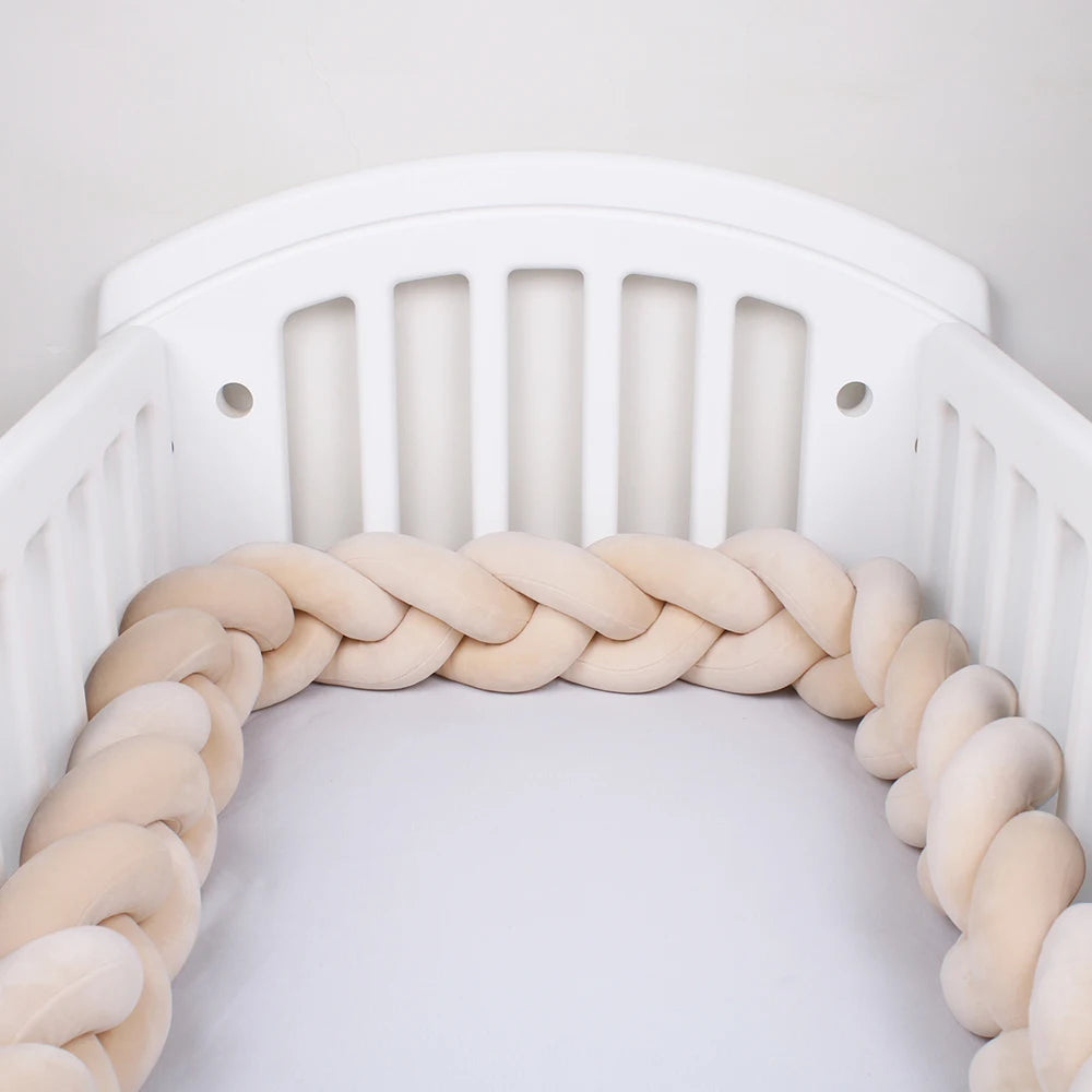 Un lit de bébé blanc avec des lattes verticales et un pare-chocs tressé entourant les bords intérieurs. Le tour de lit en peluche **Tour de lit Bébé Tressé Noué** de **BABY PREMA** est fabriqué en tissu doux et respirant et présente des brins entrelacés dans un dégradé de couleurs blanc cassé à beige. Le berceau est vide et le fond est blanc uni, assurant la sécurité de votre bébé.