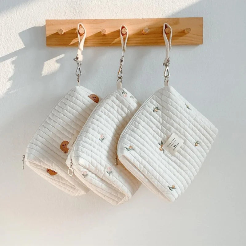Trois Sac de Couches Rangement Bébé avec des motifs de feuilles et de panaches accrochés à des piquets en bois contre un mur blanc par BABY PREMA.