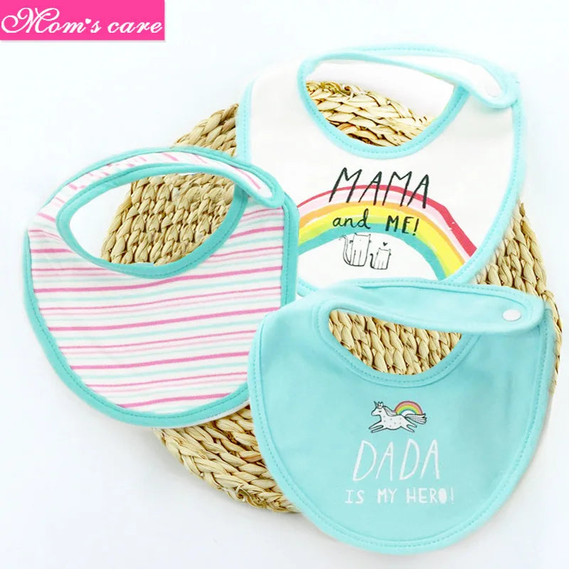 Trois **Lot de 3 pièces Bavoirs Bébé** disposés sur un tapis tissé. Le premier bavoir a des rayures roses, le second porte l'inscription « MAMA and ME ! » avec un graphique arc-en-ciel, et le troisième est turquoise avec une licorne et le texte "DADA is MY HERO !" Fabriqué en coton de haute qualité, le logo "**BABY-PREMA**" est visible dans le coin supérieur gauche.