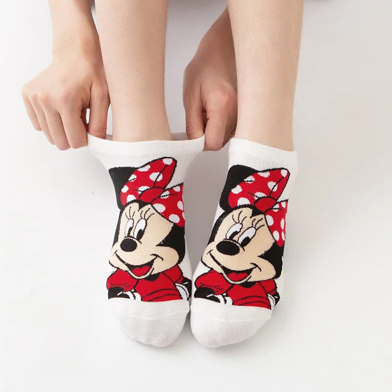 Une paire de pieds arborant des chaussettes bateau blanches avec un délicieux motif de Minnie Mouse, avec son visage, un nœud rouge à pois blancs et une robe rouge. Les mains de la personne ajustent ces Paire Chaussettes Coton Coréennes BABY PREMA sur un fond uni blanc.