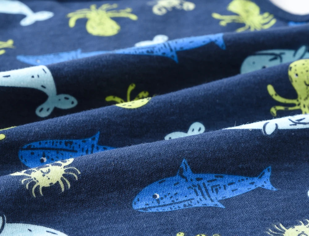Gros plan d'un tissu bleu présentant un motif ludique sur le thème marin, comprenant des baleines, des requins, des poulpes et des crabes ressemblant à des dessins animés dans différentes nuances de bleu, vert et jaune. Les légers plis du tissu mettent en valeur ses détails et sa texture, parfaits pour créer un ensemble de baby shower ou un Lot de 3 Bodies bébé de BABY PREMA.