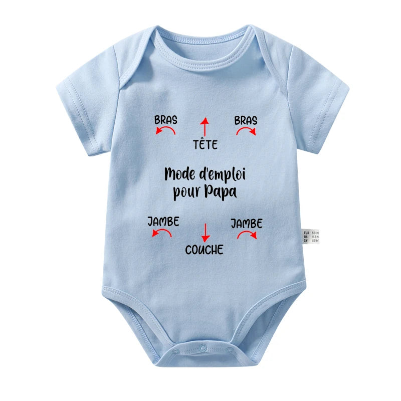 Une combinaison en coton bleu clair, parfaite pour les journées chaudes, comporte un texte en français avec des instructions pour habiller un bébé. Des flèches et des étiquettes indiquent où placer les bras (Bras), la tête (Tête), les jambes (Jambe) et la couche (Couche), avec le titre « Mode d'emploi pour Papa ». Il s'agit du Body Coton Imprimé de BABY-PREMA.