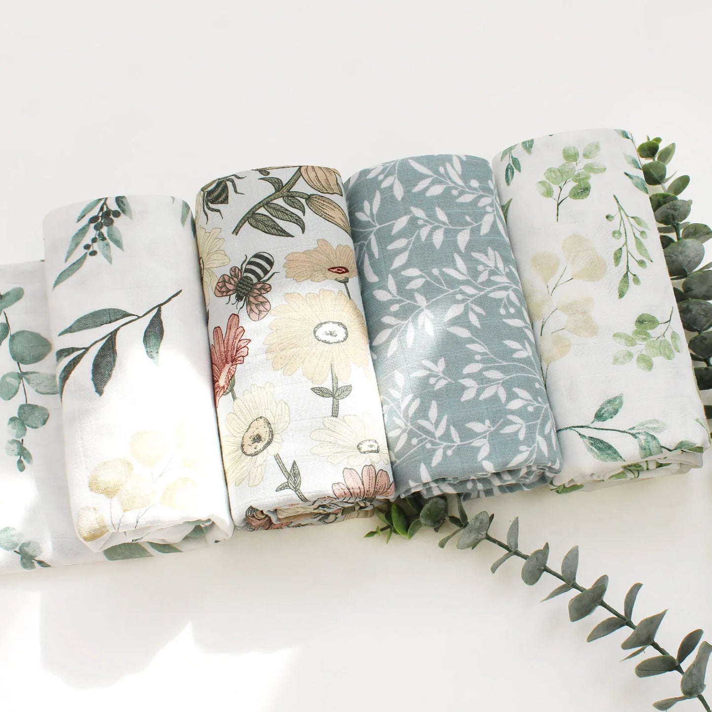 Une collection d'élégants rouleaux de tissu aux motifs floraux et botaniques, présentés avec goût avec un brin de verdure fraîche pour compléter les motifs naturels, idéal pour créer la Couverture Bébé en Coton et Bambou Bio de BABY PREMA.