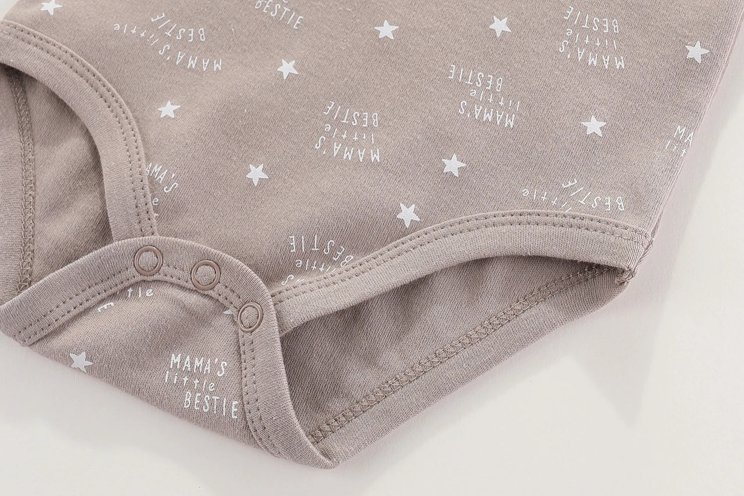 Gros plan d'une combinaison beige pour bébé avec des motifs d'étoiles blanches et "MAMA'S Little BESTIE" imprimé à plusieurs reprises. Cette adorable pièce BABY PREMA Lot 6 Body Coton Bébé Manches Courtes comporte des fermetures à pression en bas, parfaites pour votre bébé.