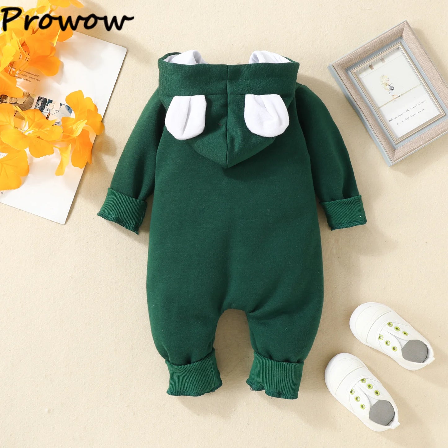 Une combinaison verte pour bébé avec une capuche ornée d'oreilles d'ours blanches repose sur une surface beige. À côté de la combinaison se trouvent une paire de petites chaussures de bébé blanches avec des boutons verts et une photo encadrée d'une girafe. Des feuilles artificielles jaunes sont également placées à proximité, créant le cadre idéal pour BABY PREMA Combinaison pour Nouveau-nés.