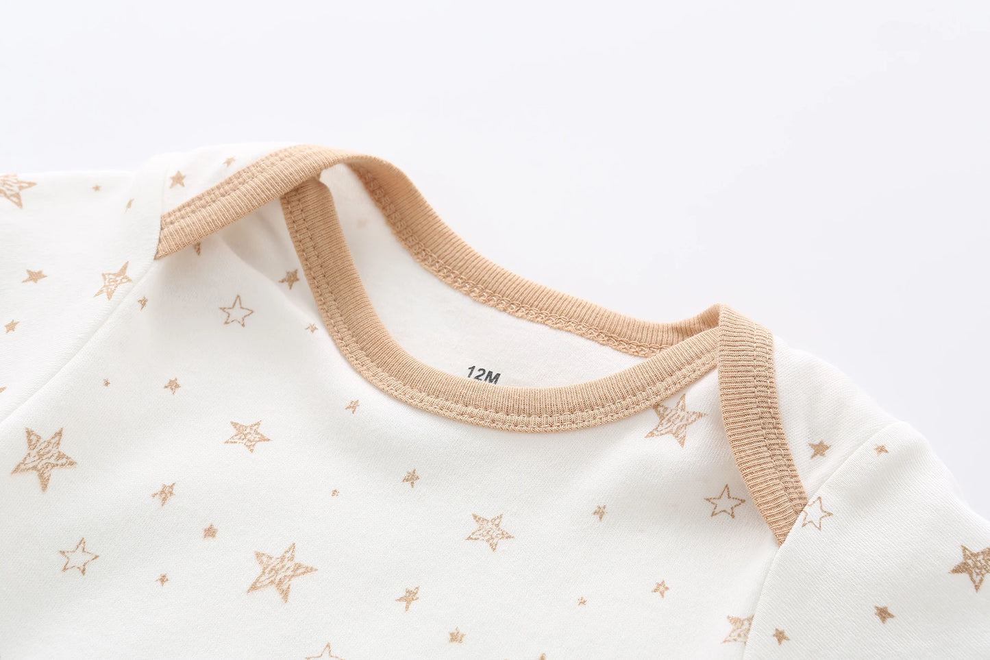 Une combinaison blanche pour bébé étiquetée « 12M » présente des motifs d'étoiles marron clair et une bordure beige autour de l'encolure. Cette adorable pièce, parfaite pour un trousseau de naissance, est confectionnée dans un coton doux et respirant pour assurer le confort de votre bébé. Il fait partie du Lot de 5 Bodys Bébé | Collection Coton Agréables & conforts par BABY PREMA.