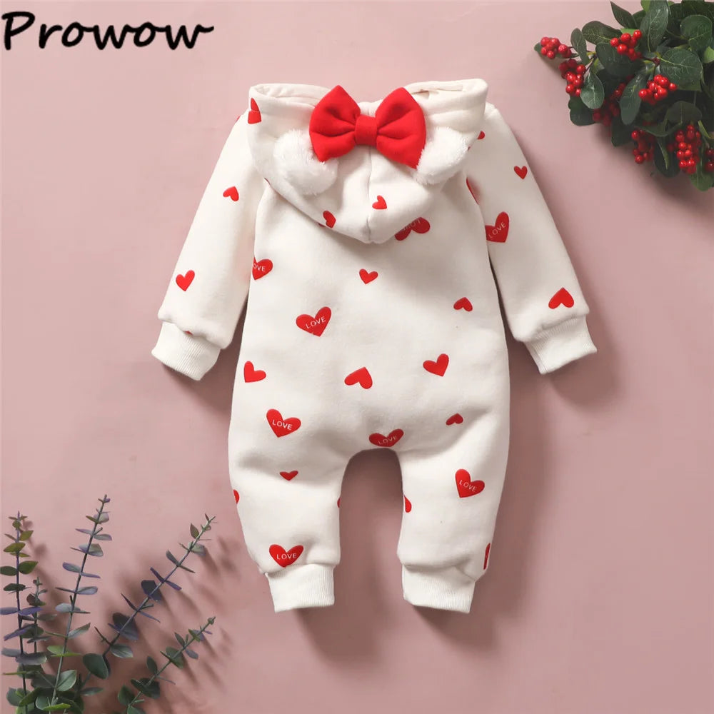 Une Combinaison pour Nouveau-nés avec une capuche ornée de motifs cœurs rouges et un grand nœud rouge au dos. Cette tenue blanche, qui rappelle les vêtements d'hiver pour bébé, est posée à plat, entourée de quelques brins verts et baies rouges. Le mot « BABY PREMA » est imprimé dans le coin supérieur gauche.