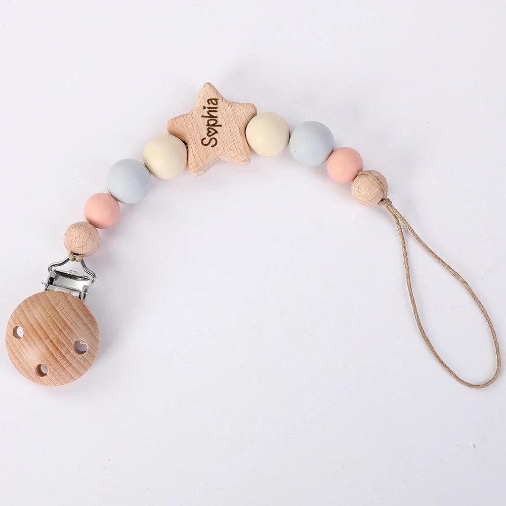 Une attache tétine bébé en bois personnalisée avec le prénom "sylvana" gravé sur une perle en forme d'étoile, comportant une Attache Sucette Bébé Personnalisé BABY-PREMA de perles rondes en bois naturel et de couleur pastel, fixées sur une attache en métal.