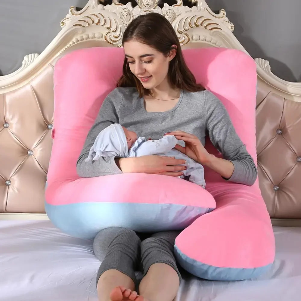 Une femme est assise sur un lit, berçant un bébé dans ses bras. Adossée à un grand Coussin de Maternité & Allaitement rose et bleu de BABY PREMA avec une tête de lit ornée derrière elle, elle est vêtue d'une tenue d'intérieur grise. La femme et le bébé semblent calmes et satisfaits, enveloppés de soutien et de confort.