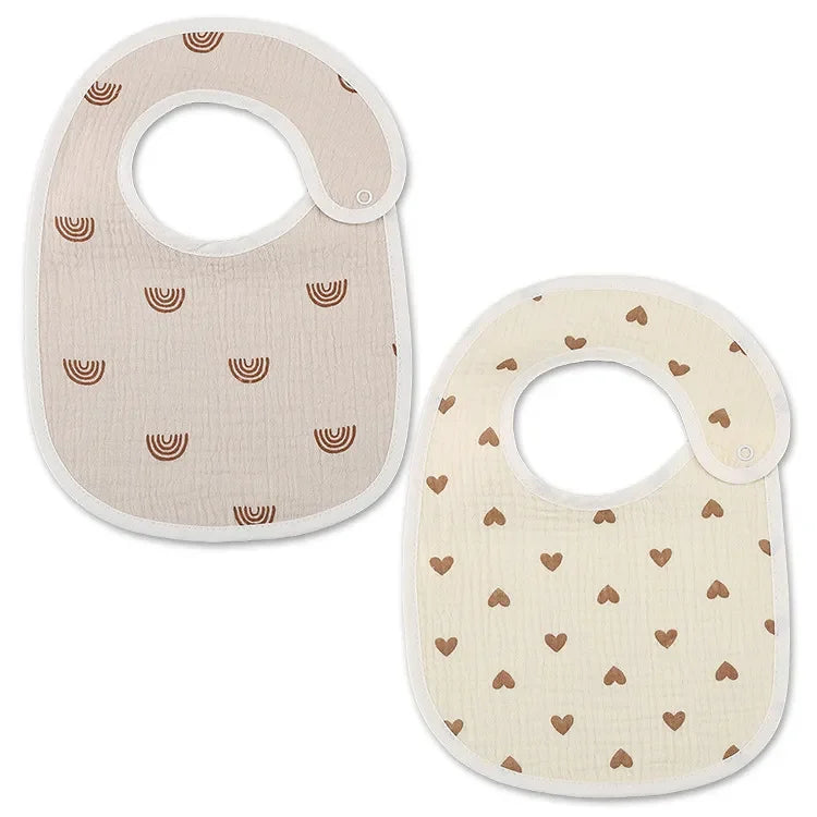 Deux bavoirs pour bébé sont représentés, l'un en beige avec des arcs marron et l'autre en blanc cassé orné de cœurs marron. Les deux Bavoirs Motifs Bébé 2 Pièces de BABY-PREMA ont une fermeture à col arrondi avec bouton pression, alliant fonctionnalité et design charmant.