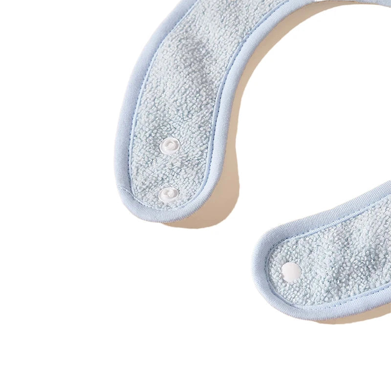 Vue rapprochée d'un article en tissu bleu clair, éventuellement un bavoir pour bébé ou un accessoire vestimentaire avec un motif bandana, comportant deux boutons-pression à chaque extrémité. Le tissu semble avoir une surface douce, texturée et rembourrée. L'article est disposé sur un fond blanc uni. Cet article fait partie des Bavoirs Bébé Lot de 4 Pièces de BABY-PREMA.