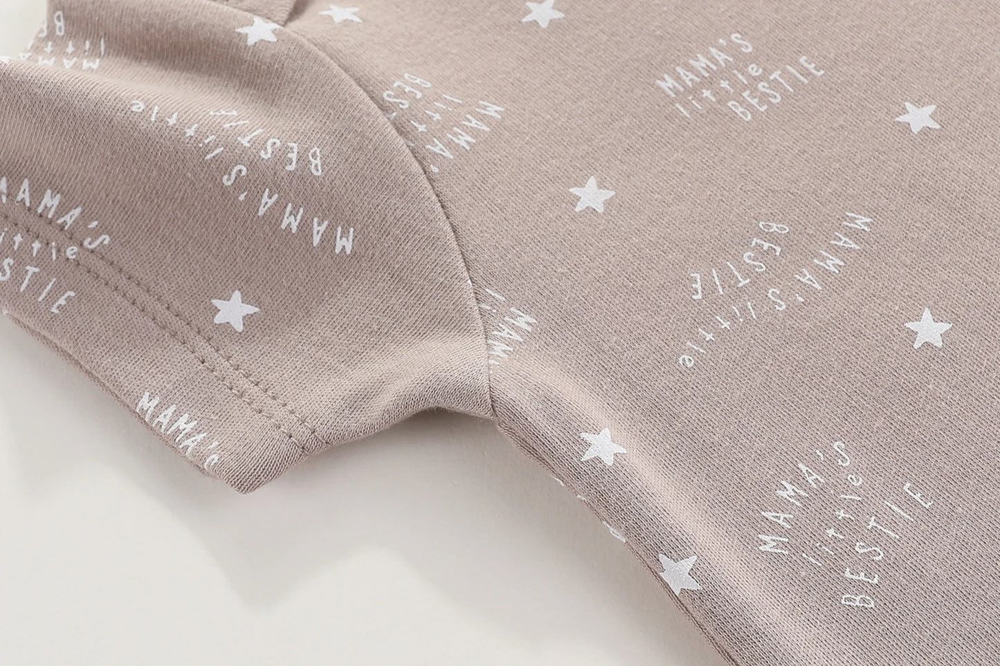 Gros plan sur un tissu marron clair avec un texte imprimé en blanc indiquant « La petite meilleure amie de Mama » et de petits motifs d'étoiles blanches. La texture de ce tissu en coton à manches courtes avec coutures apparentes met en valeur sa qualité, complétée par des boutons-pression pratiques pour un habillage facile. Il s'agit du Lot 6 Body Coton Bébé Manches Courtes de la marque BABY PREMA.