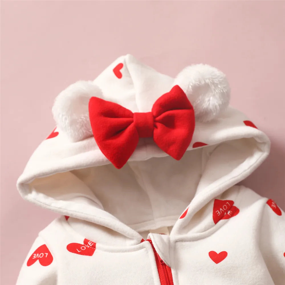 Un sweat à capuche blanc avec des motifs de coeurs rouges et une fermeture éclair rouge, parfait comme vêtement d'hiver pour bébé. La capuche comporte des oreilles d'ours en fourrure blanche et un grand nœud rouge au centre. Le mot « LOVE » est imprimé à l’intérieur de certains cœurs rouges. Le fond est rose clair. Recherchez la Combinaison pour Nouveau-nés de BABY PREMA pour la tenue d'hiver parfaite pour votre bébé !
