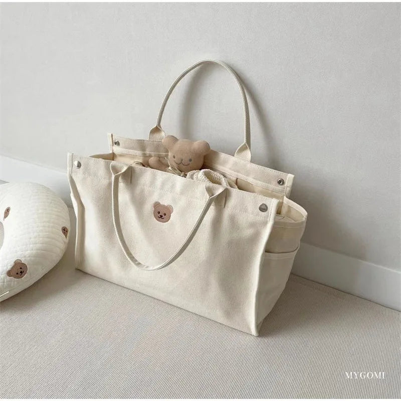 Un ours en peluche douillet jaillit d'un sac fourre-tout BABY-PREMA Sac à Langer Bébé Fourre Tout Beige à côté d'un coussin avec un motif d'ours similaire, évoquant un sentiment de minimalisme charmant et d'esthétique apaisante pour mon bébé.