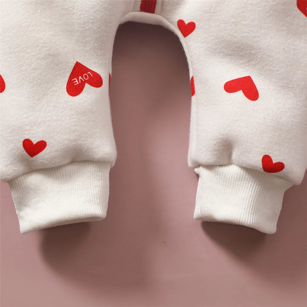 Gros plan sur la partie basse d'un pantalon bébé blanc à motifs coeurs rouges. Certains cœurs portent le mot "AMOUR" écrit à l'intérieur. Le pantalon, parfait pour l'hiver, s'affiche sur un fond rose. Le tissu en coton avec des poignets côtelés aux chevilles est visible, similaire en termes de confort à une confortable BABY PREMA Combinaison pour Nouveau-nés.