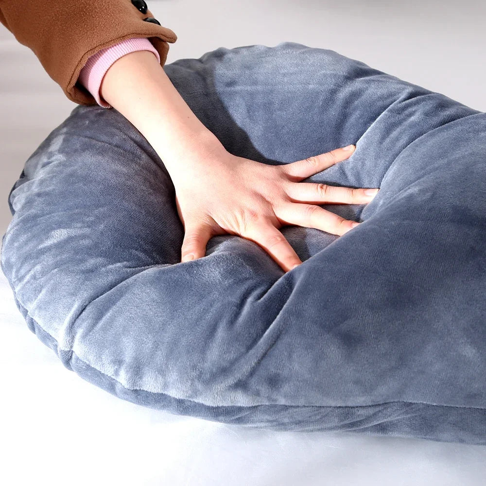 La main d'une personne appuyant sur un Coussin de Maternité & Allaitement gris en peluche de BABY PREMA, créant une empreinte. Le coussin semble doux et a une texture veloutée, offrant soutien et confort. L'individu porte un pull marron avec une manchette rose visible au poignet.