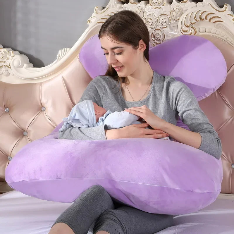 Une femme enceinte est assise sur un lit avec une tête de lit ornée, berçant un bébé enveloppé dans une couverture. Elle utilise un grand Coussin de Maternité & Allaitement violet de BABY PREMA pour son soutien et son confort. Les deux semblent détendus et confortables. La salle dégage une ambiance douce et chaleureuse.