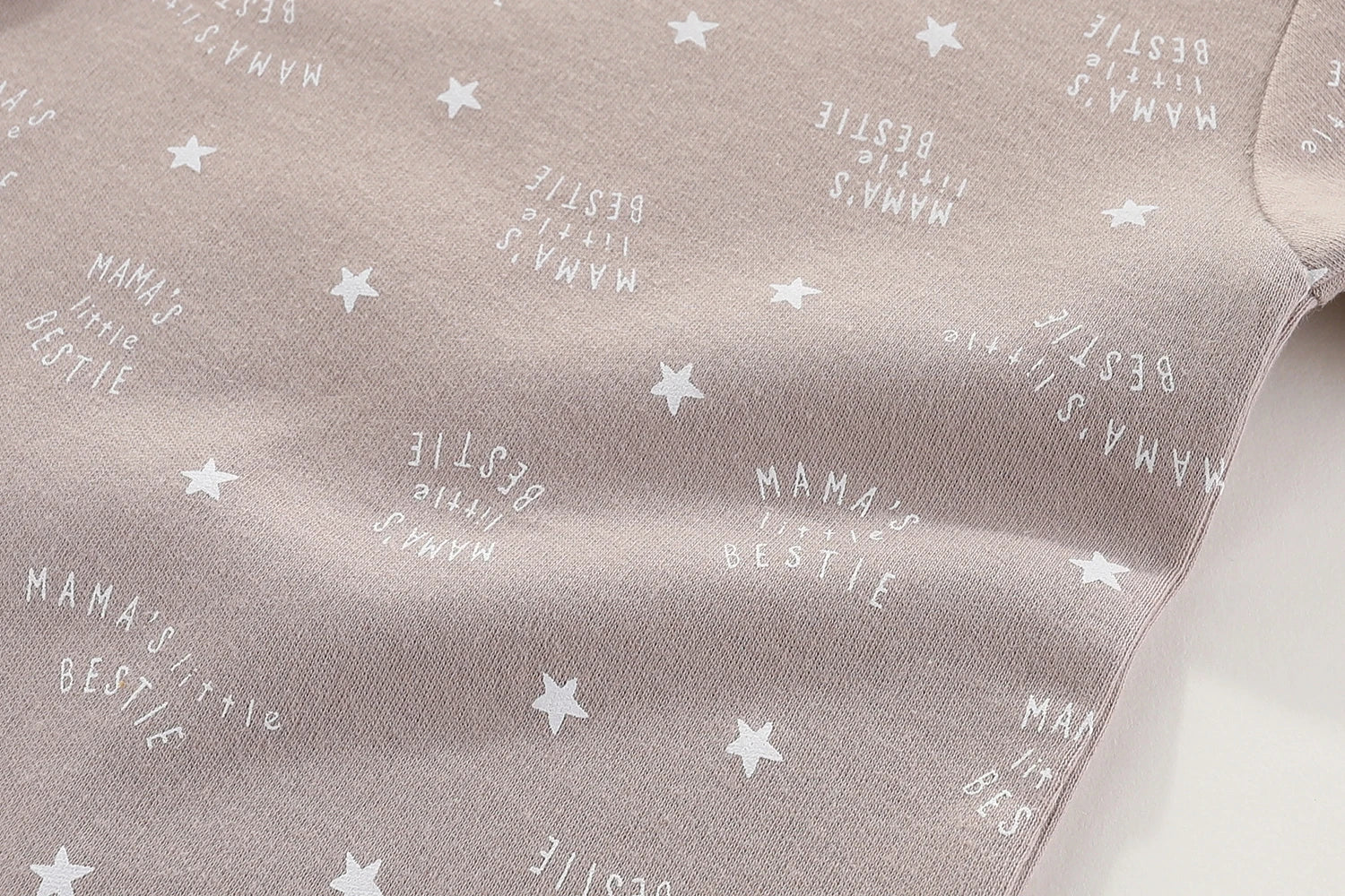 Gros plan d'un tissu gris clair avec un motif d'étoiles blanches et le texte "MAMA'S BESTIE" répété partout, avec des manches courtes et des boutons pression pour un habillage facile, mettant en valeur le Lot 6 Body Coton Bébé Manches Courtes de BABY PREMA.