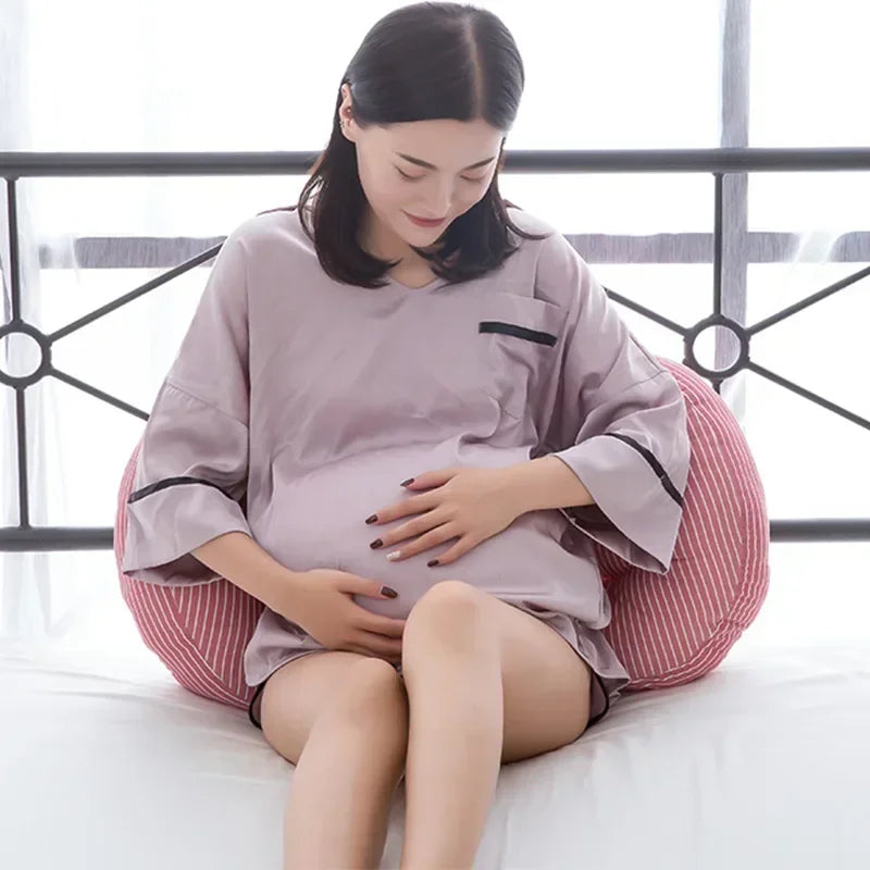 Une femme enceinte est assise sur un lit, adossée à un Oreiller de Grossesse en Coton de BABY PREMA à rayures roses. Elle porte une chemise de nuit ample gris clair et pose doucement ses mains sur son ventre. L’arrière-plan présente un cadre de lit en métal et une literie de couleur claire.