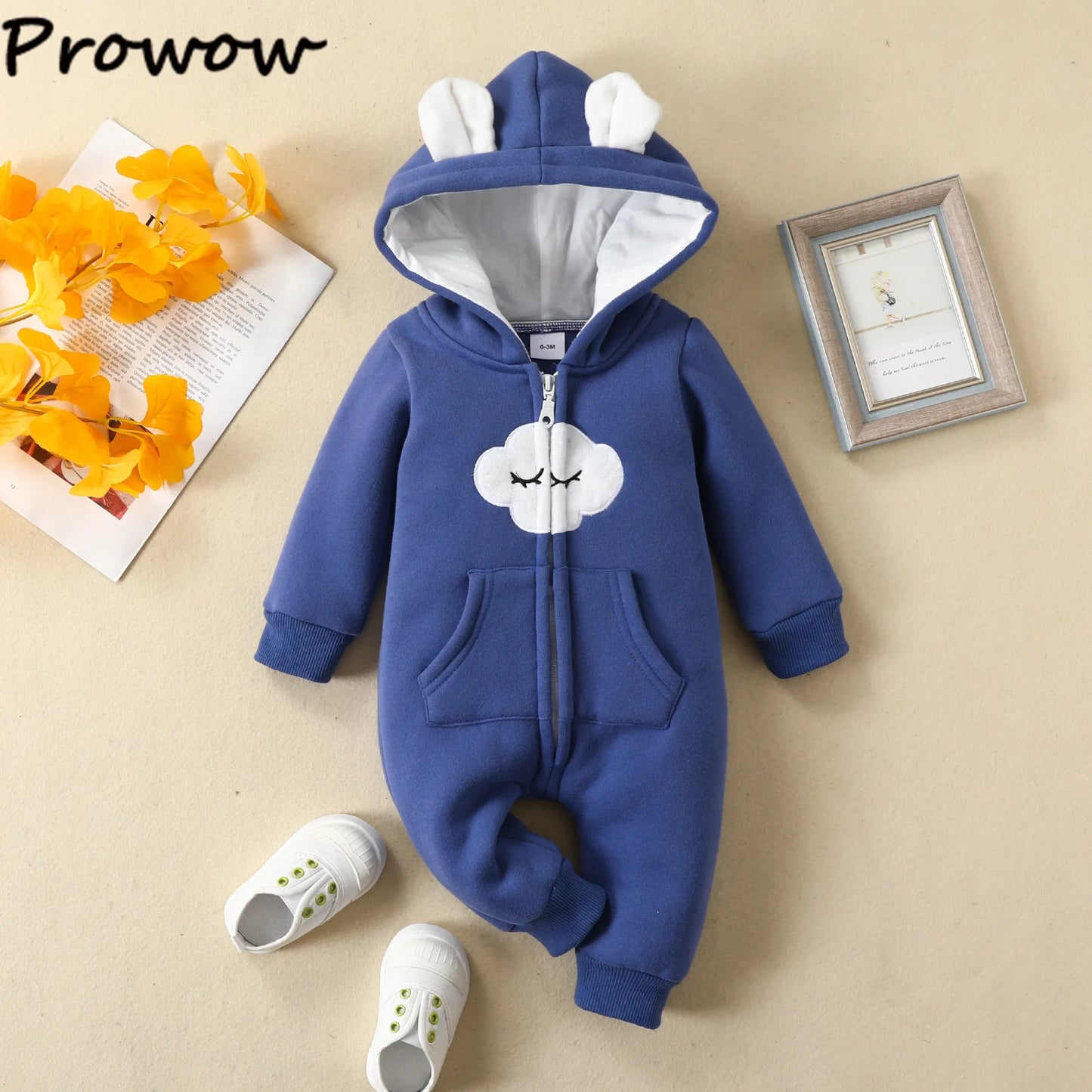 Une combinaison bleue pour bébé avec une capuche avec des oreilles d'ours et un joli motif nuage sur le devant est disposée sur une surface de couleur claire. À côté se trouvent des chaussures de bébé blanches avec des accents verts, une photo encadrée et un bouquet de fleurs jaunes. Cette adorable Combinaison à Capuche Barboteuse pour nouveau-né de la marque "BABY PREMA" est tout simplement charmante.