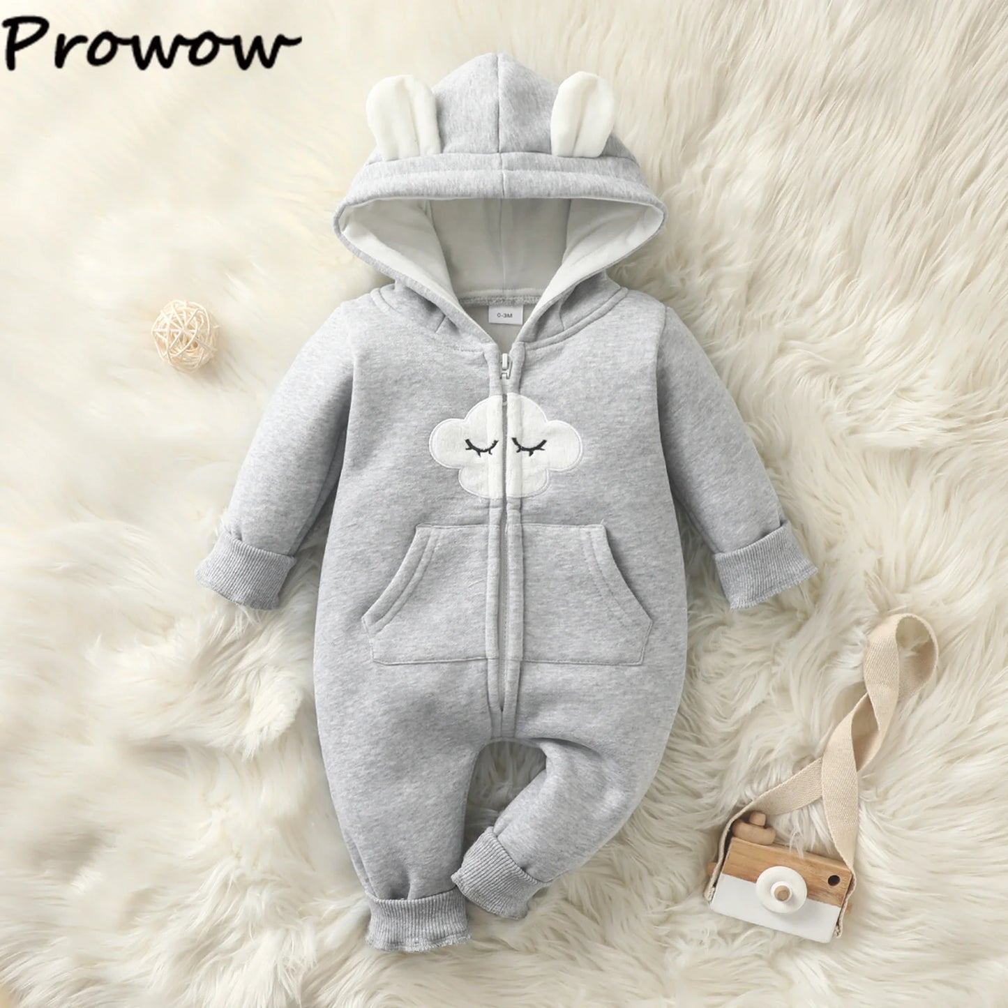 Une Combinaison pour Nouveau-nés grise douillette de BABY PREMA avec une capuche avec des oreilles blanches en forme d'ours et un joli motif de nuage endormi sur la poitrine. L'image comprend également un petit appareil photo en bois avec une sangle beige et une pelote de laine, le tout placé sur un fond doux, de couleur crème et moelleux : des vêtements d'hiver parfaits pour bébé.