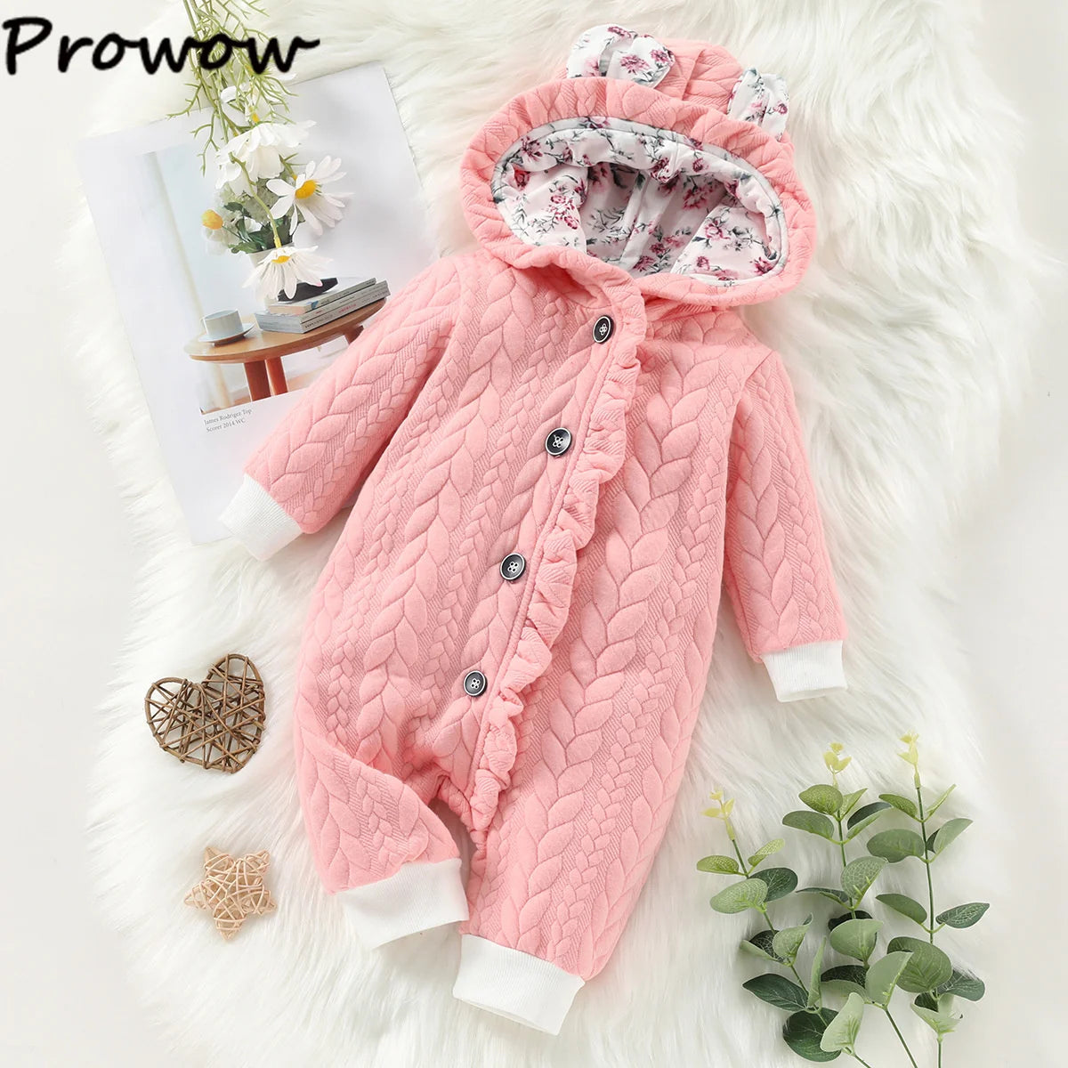 Une Combinaison pour Nouveau-nés rose et matelassée de BABY PREMA avec une capuche ornée d'une doublure en tissu floral et d'un bord volanté est disposée sur une surface blanche. Autour de lui se trouvent une plante, une décoration en forme de cœur tissée et un magazine ouvert présentant des vêtements d'hiver pour bébé ainsi qu'un adorable petit objet en forme d'étoile.