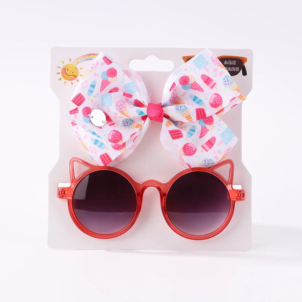 Ensemble d'accessoires pour enfants comprenant des lunettes de soleil pour bébé rondes à monture rouge avec des détails d'oreilles de chat et un nœud coloré à motifs de crème glacée, présentés sur un support en carton blanc.
BABY PREMA Ensemble d'Accessoires Bébé | Lunettes de Cheveux pour Bébé.