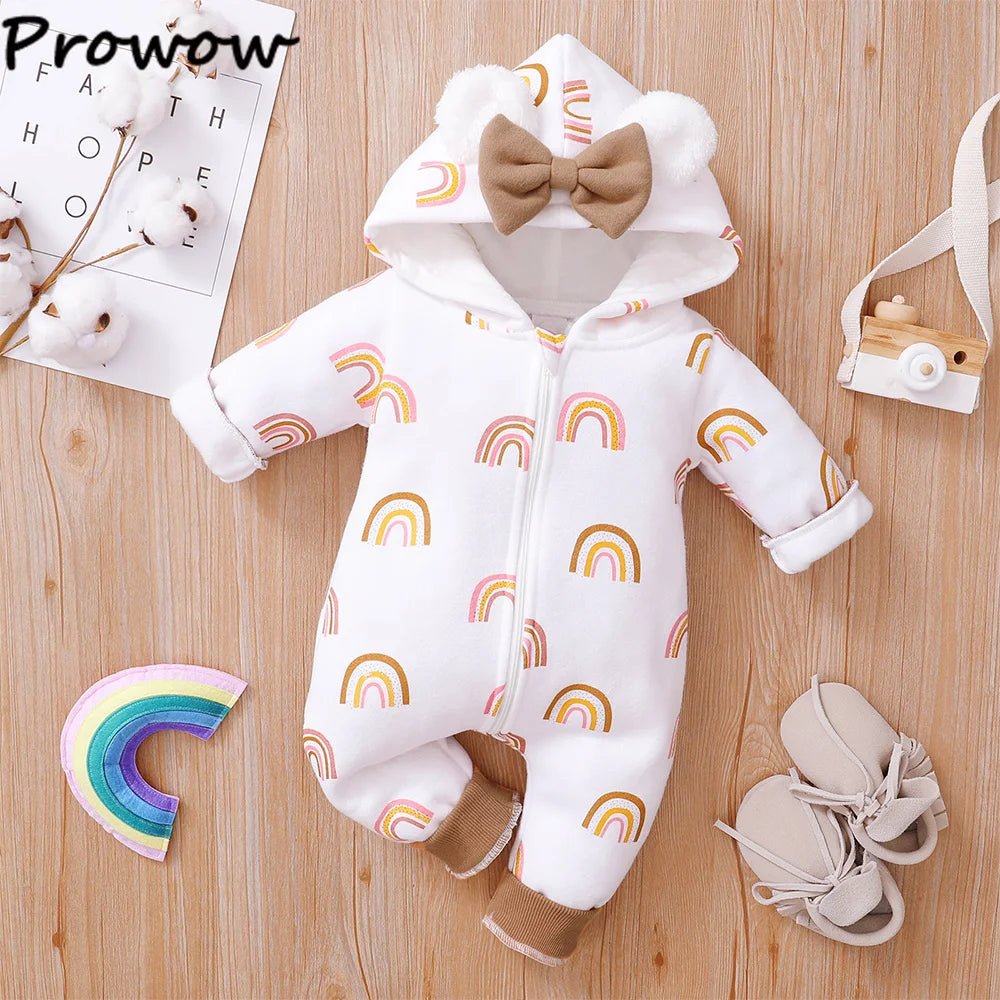 Une Combinaison pour Nouveau-nés BABY PREMA aux imprimés arc-en-ciel repose sur une surface en bois. La tenue blanche comprend une capuche avec des oreilles d'ours et un nœud marron, parfaite pour l'hiver. Les articles d'accompagnement comprennent des chaussures de bébé, un appareil photo jouet en bois, un jouet en forme d'arc-en-ciel et des fleurs en coton avec une œuvre d'art en arrière-plan.