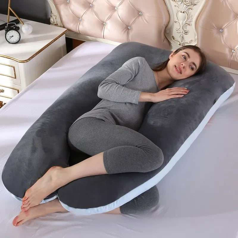 Une femme est allongée sur le côté sur un lit, câlinant un grand Coussin de Maternité & Allaitement en forme de U de BABY PREMA. Vêtue d'un pyjama gris, elle apparaît confortable et détendue. Le lit a une tête de lit capitonnée et une table de chevet avec une horloge dessus, offrant soutien et confort à la femme enceinte.