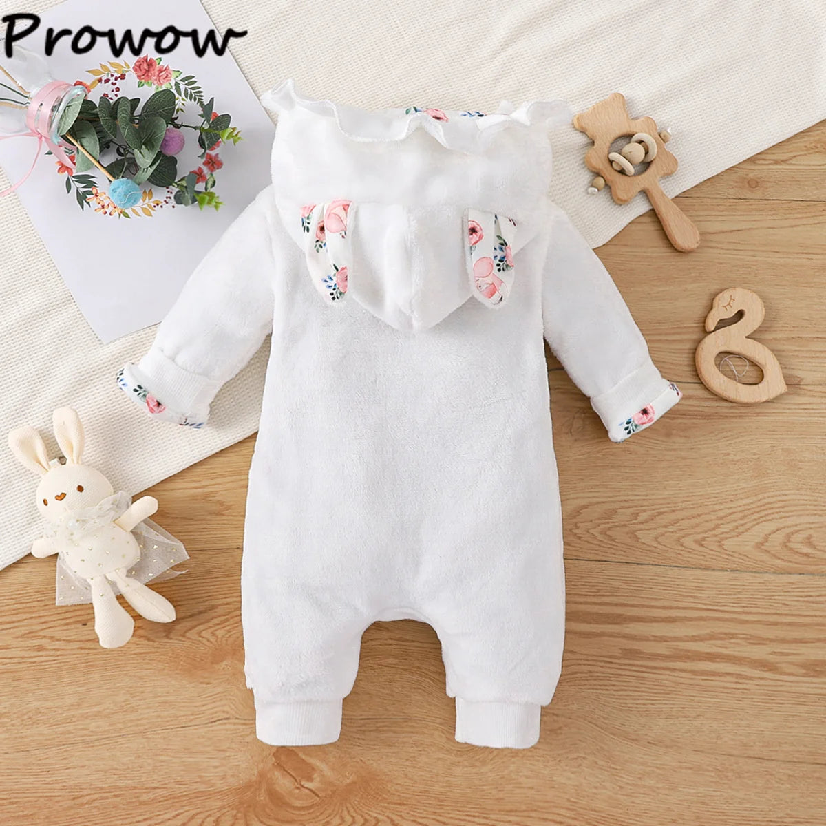 Une combinaison pour bébé avec des oreilles d'animaux et une capuche fleurie repose sur une surface en bois. Autour de lui se trouvent un lapin en tissu, un hochet en bois et une composition florale dans le coin, sous un éclairage doux. Parfaite pour l’hiver, cette Combinaison pour Nouveau-nés est douillette et adorable. Le nom « BABY PREMA » apparaît dans le coin supérieur gauche de l'image.