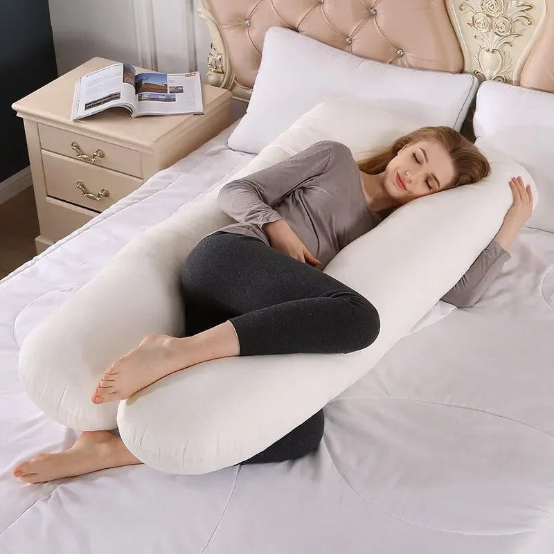 Une femme enceinte aux cheveux longs est allongée sur un lit blanc, serrant dans ses bras un grand Coussin de Maternité & Allaitement en forme de U de BABY PREMA pour soutien et confort. Elle porte une chemise grise à manches longues et des leggings noirs. À côté du lit, il y a une table de nuit en bois avec un livre ouvert et des lunettes dessus.