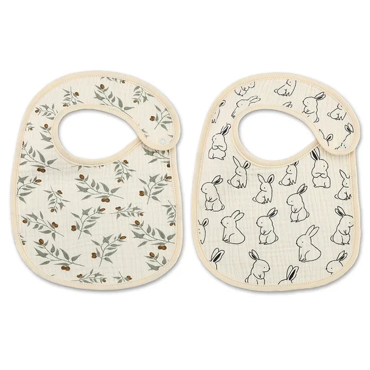 Deux bavoirs pour bébé avec trous au col rond et fermetures boutonnées. Un bavoir présente un motif feuillu ; l'autre a un imprimé lapin. Les deux sont des Bavoirs Motifs Bébé 2 Pièces de couleur claire de BABY-PREMA.
