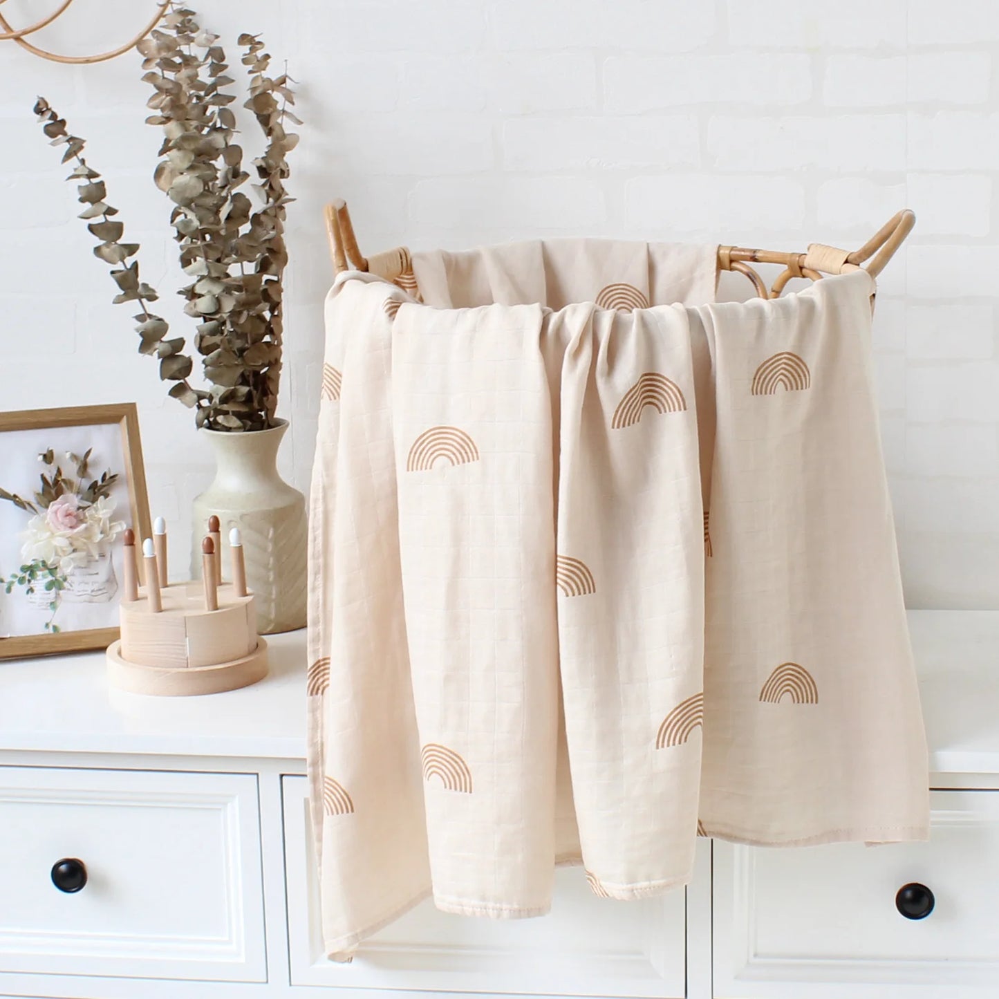 Un élégant panier à linge beige avec des motifs semi-circulaires se dresse dans une pièce blanche et épurée avec un vase d'eucalyptus, un cadre photo et des objets décoratifs dont la Couverture Bébé en Coton et Bambou Bio de BABY PREMA.