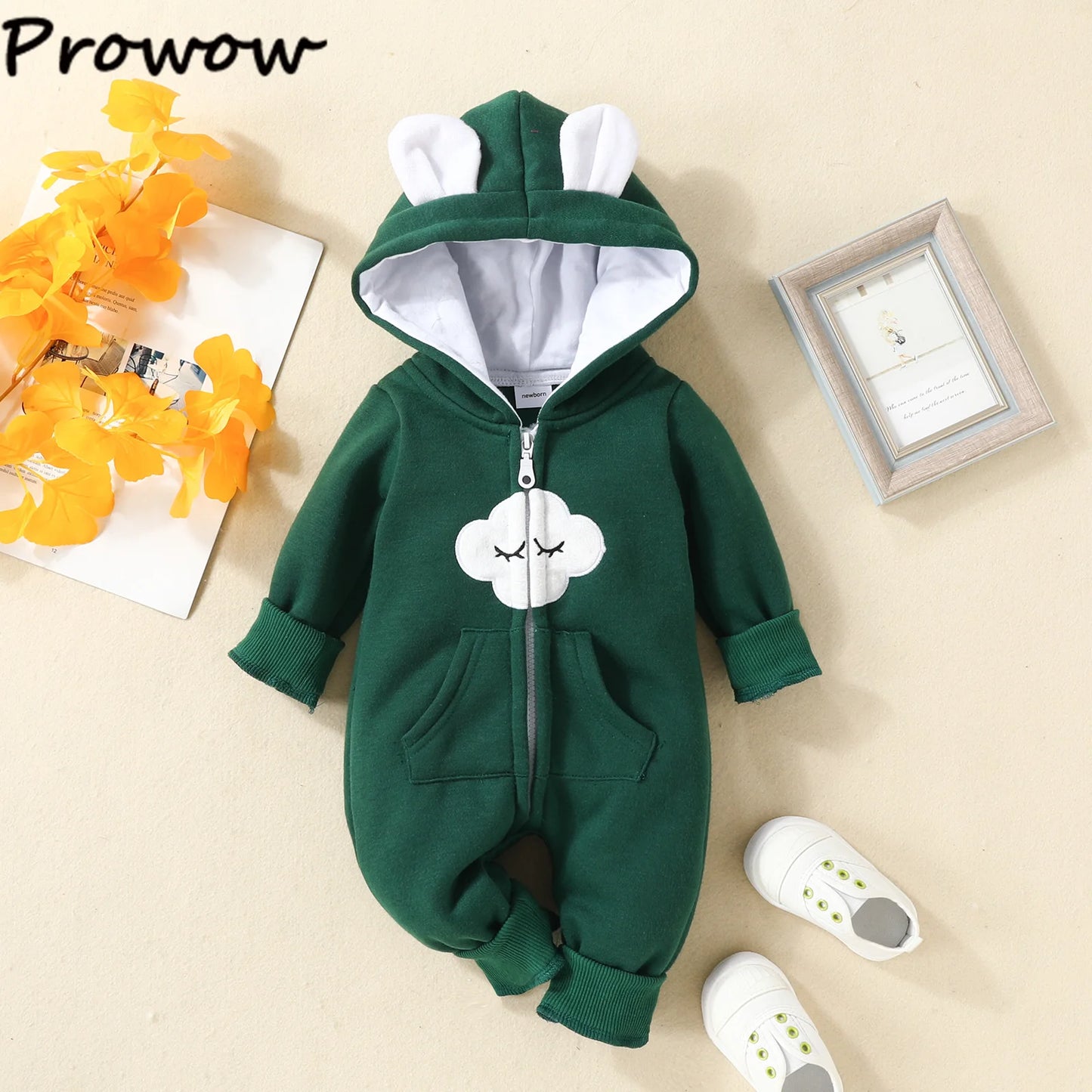 Une Combinaison pour Nouveau-nés verte avec une fermeture éclair, dotée d'une capuche avec des oreilles blanches et d'un nuage blanc avec un visage endormi sur la poitrine, parfaite comme vêtements d'hiver pour bébé de BABY PREMA. La combinaison est disposée à plat sur une surface beige à côté d'une paire de chaussures de bébé blanches, d'un cadre photo et de fleurs décoratives orange.