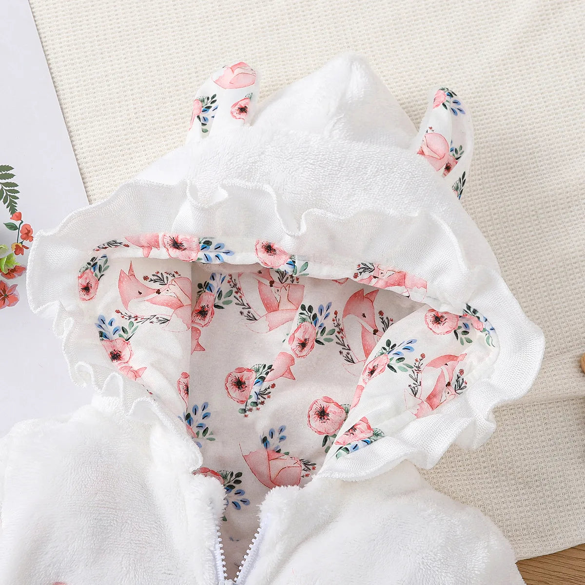 Un gros plan d'une veste à capuche pour bébé montre la doublure intérieure, présentant un motif floral avec des fleurs roses et des feuilles vertes sur fond blanc. La partie extérieure de la capuche est blanche avec une bordure à volants et deux petites oreilles en tissu sur le dessus, parfaite pour être associée à n'importe quelle Combinaison pour Nouveau-nés de BABY PREMA.
