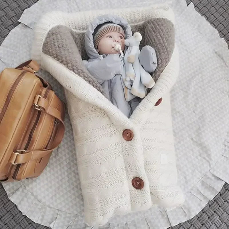 Un paquet de joie douillet : un bébé content, confortablement enveloppé dans une couverture tricotée de couleur crème avec des accessoires pour bébé, complété par un élégant sac en cuir marron à côté. Couverture de Poussette Pour Bébé de BABY PREMA.