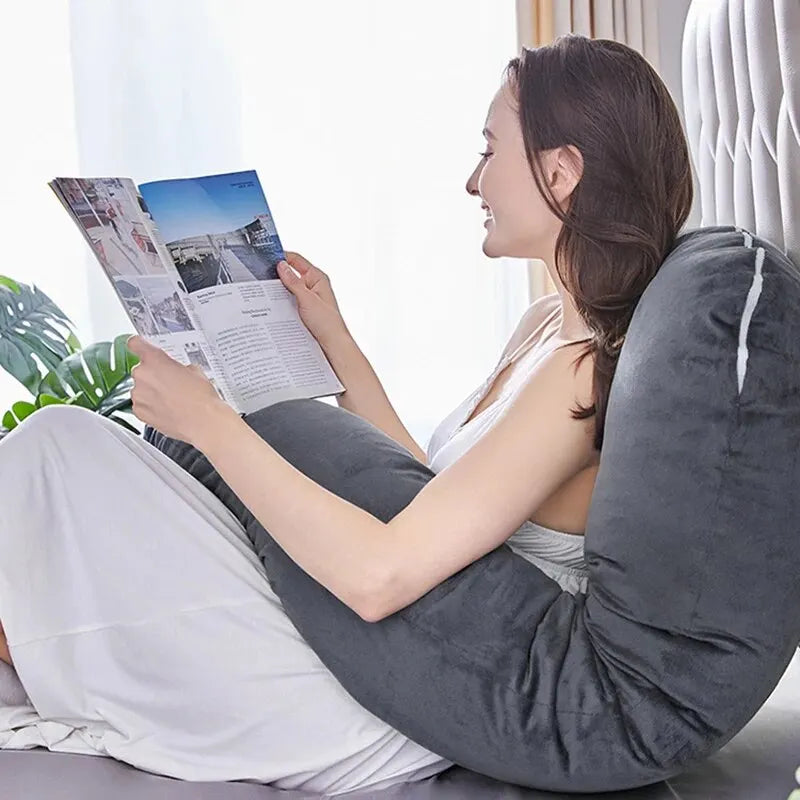 Une femme est assise confortablement dans son lit, soutenue par un grand Coussin Oreiller Femmes Enceinte gris de BABY PREMA, tout en lisant un magazine. Elle semble détendue et sourit légèrement. Il y a une plante verte visible à l’arrière-plan, avec la douce lumière du soleil filtrant à travers des rideaux de couleur claire.